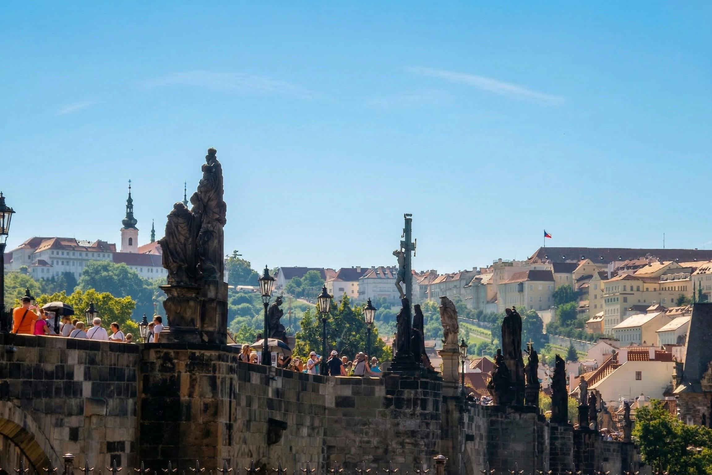 A historic bridge with statues, people walking, and a skyline of buildings and hills under a clear blue sky.