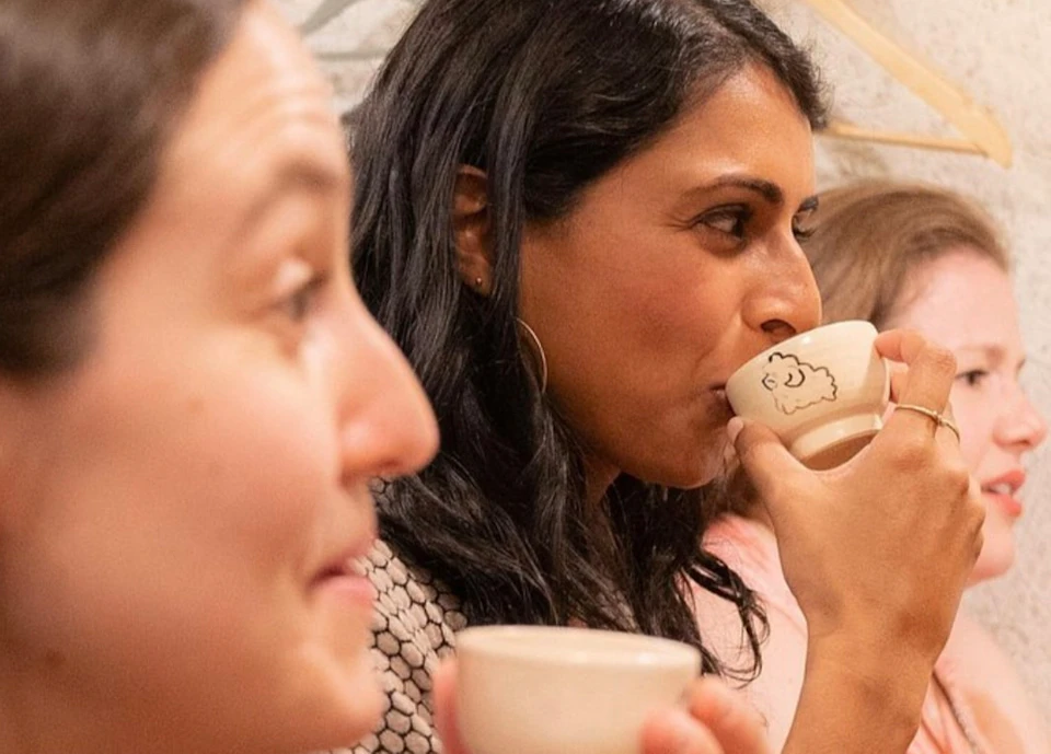 Three people sitting indoors, drinking from small cups in a cozy setting with hooks and hangers on the wall behind them.