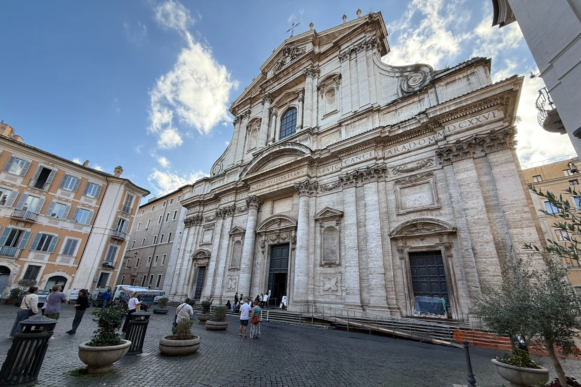 A large ornate stone building with columns and detailed carvings, surrounded by people and trees under a partly cloudy sky.