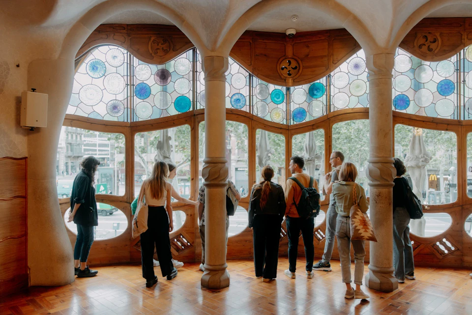 A group of people observing intricate stained glass windows and wooden details in a brightly lit interior space.