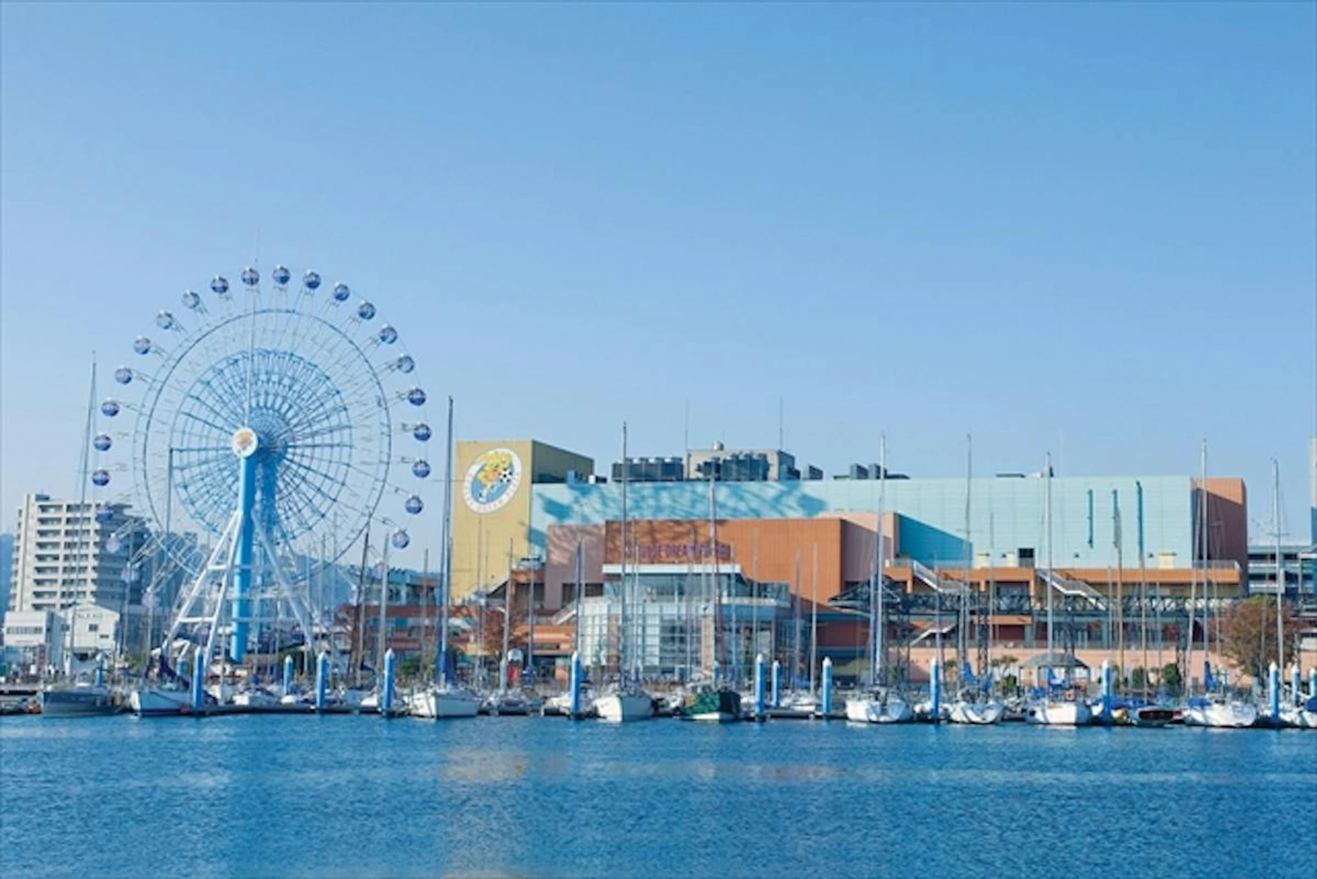 A waterfront scene featuring a Ferris wheel, a building complex, and a marina with docked sailboats under a clear blue sky.