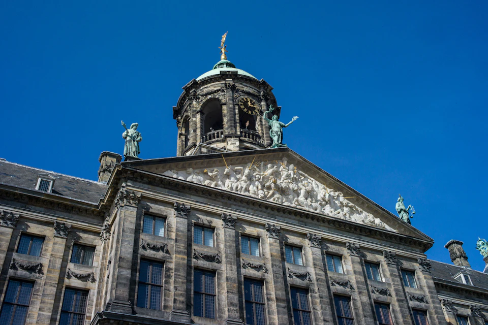 Ornate building facade with statues and relief carvings, topped by a clock tower and dome against a clear blue sky.