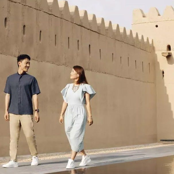 Zwei Personen gehen an einer beigen, zinnenbewehrten Mauer mit kleinen Fenstern entlang; die eine schaut die andere lächelnd an.