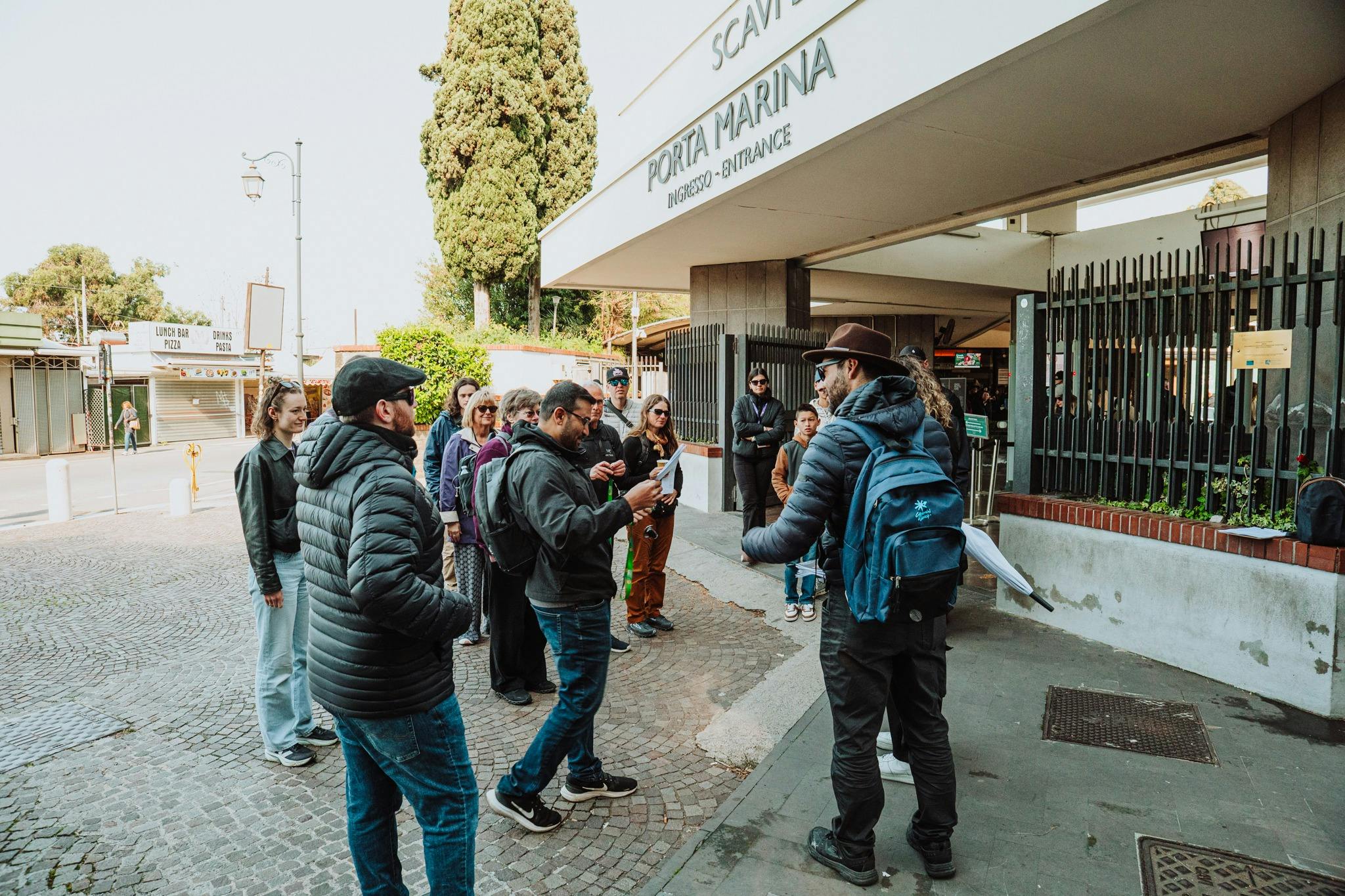 A group of people gathered near a building labeled 'Porta Marina' on a sunny day, with trees and pavement visible around.