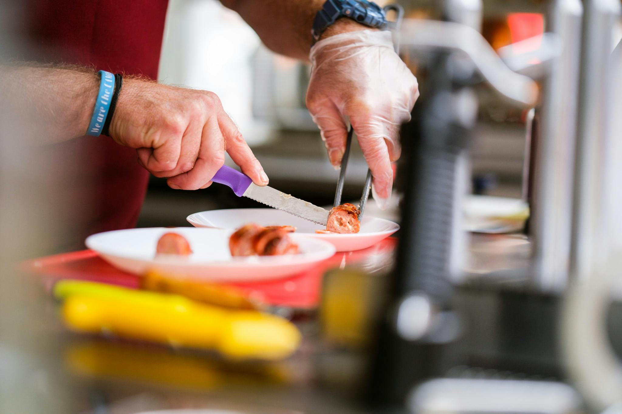 Osoba v rukavicích krájí klobásy na talíři nožem a vidličkou v kuchyňském prostředí.