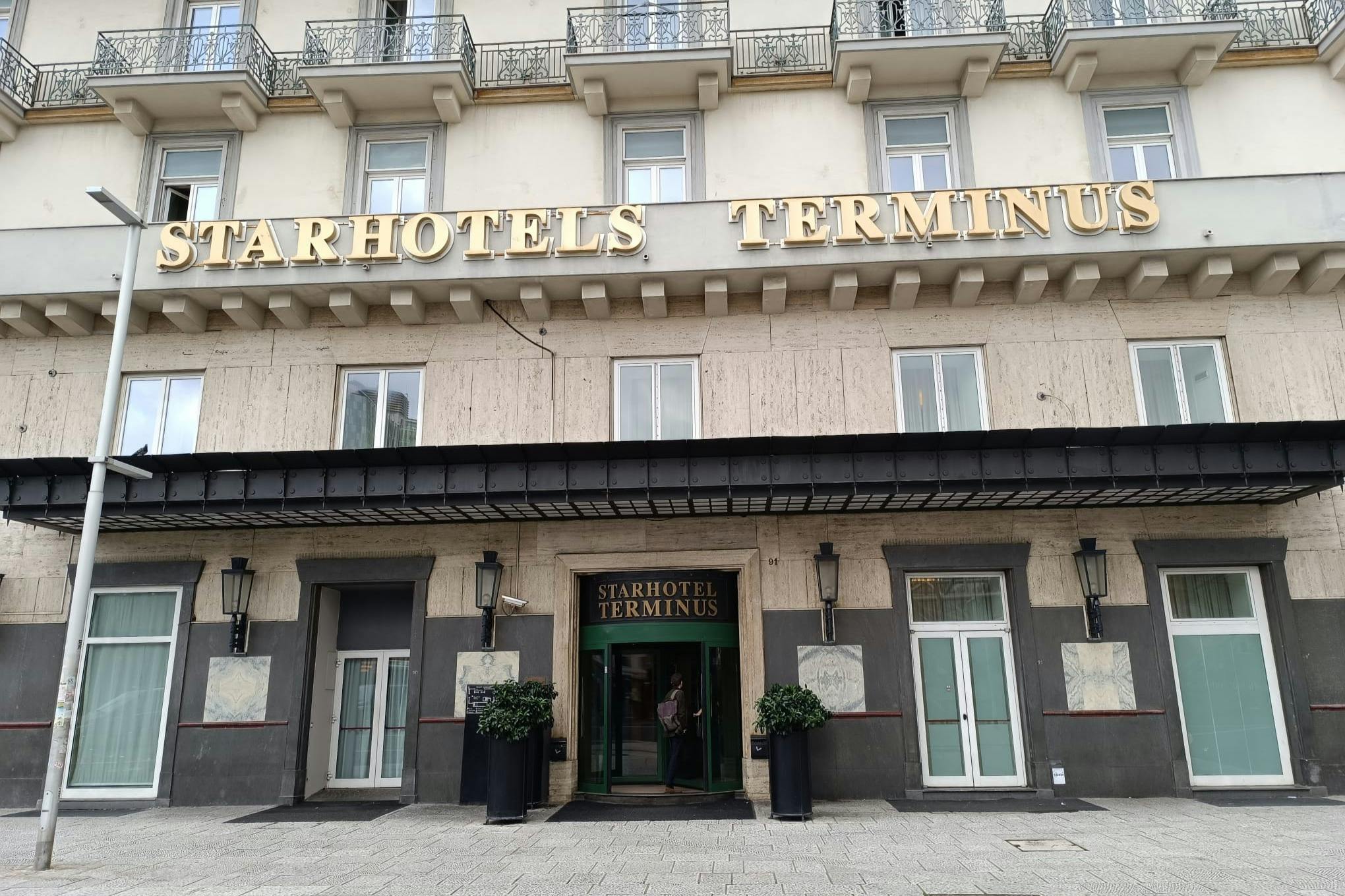 Façade du Starhotels Terminus avec une entrée centrale flanquée de grandes fenêtres et de plantes en pot, située sous un bâtiment beige avec des balcons.
