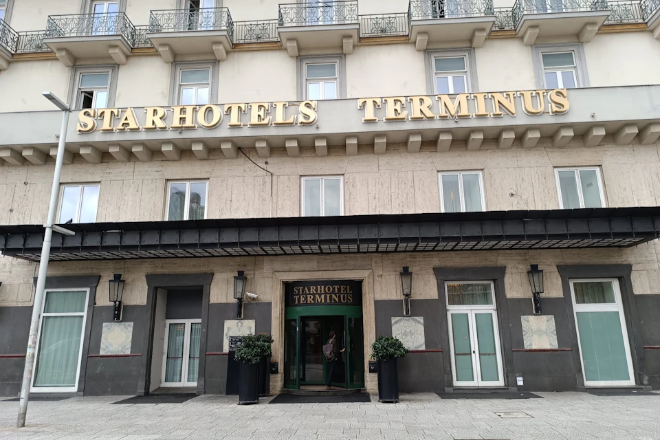 Facade of Starhotels Terminus with a central entrance flanked by large windows and potted plants, set under a beige building with balconies.