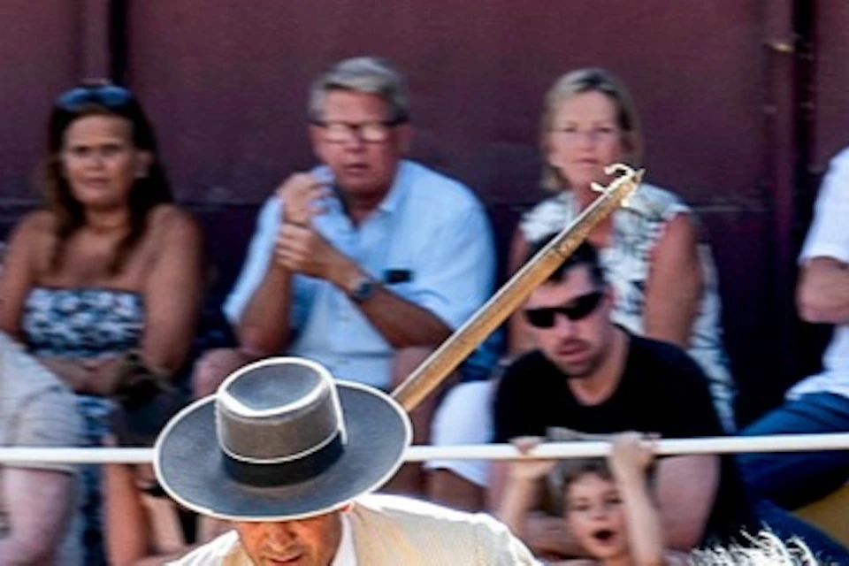 A horseman in traditional attire performs with a long pole on a galloping white horse in an arena with spectators.