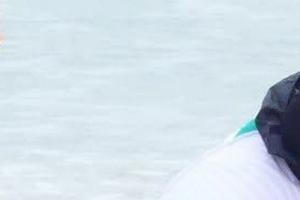 A man in a green shirt practices surfing poses on a blue surfboard on the beach while another man in a white shirt instructs him.