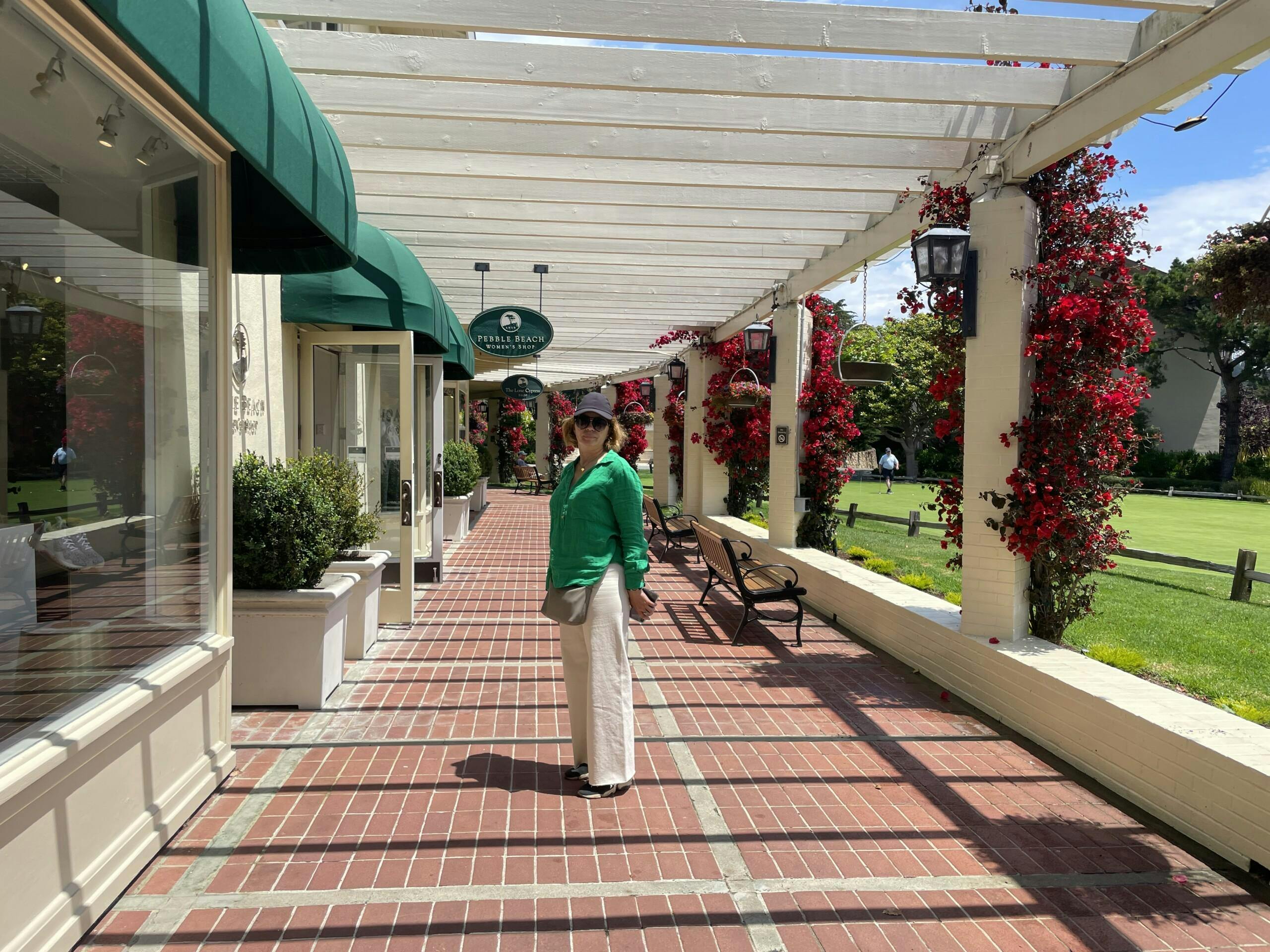 Ένα άτομο στέκεται σε έναν τούβλινο διάδρομο κάτω από μια πέργκολα με πράσινο και κόκκινα λουλούδια στο παρασκήνιο.