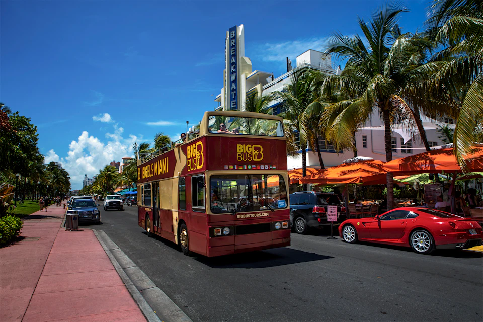 A double-decker tour bus drives down a street lined with palm trees and outdoor seating under orange umbrellas.