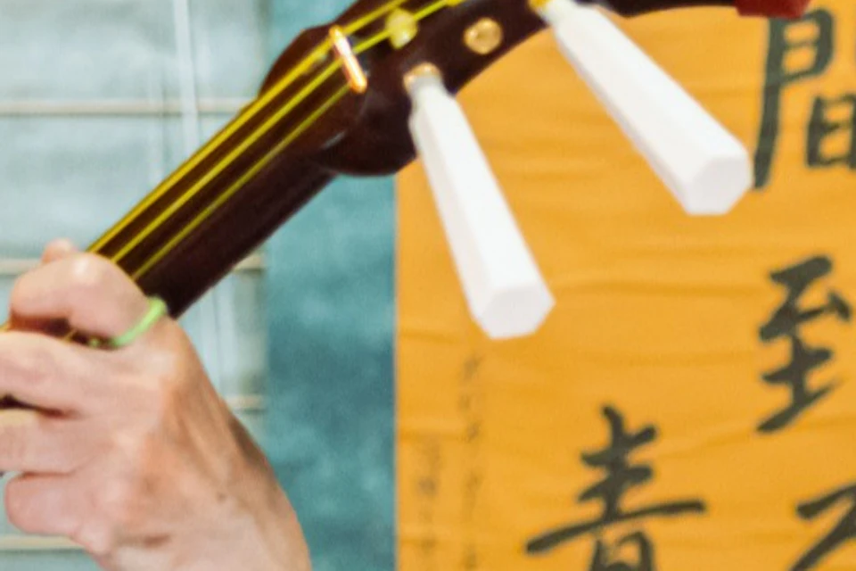 Two women in traditional attire perform on shamisen instruments in an indoor setting with a brick wall and Japanese calligraphy.