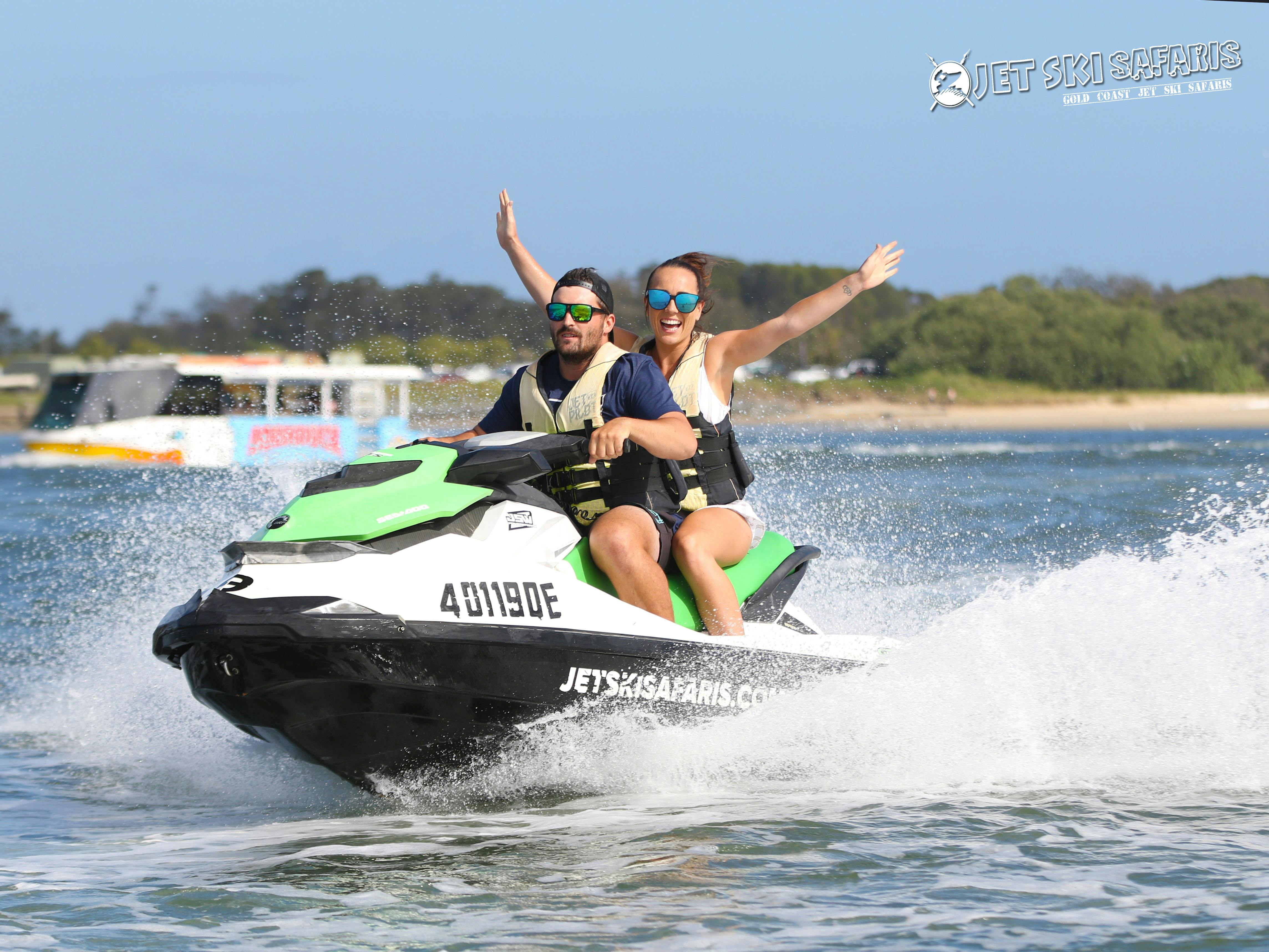 两个穿着救生衣的人在水上骑摩托艇。 后面的女人举起了手臂。 后面是一艘船和树木。