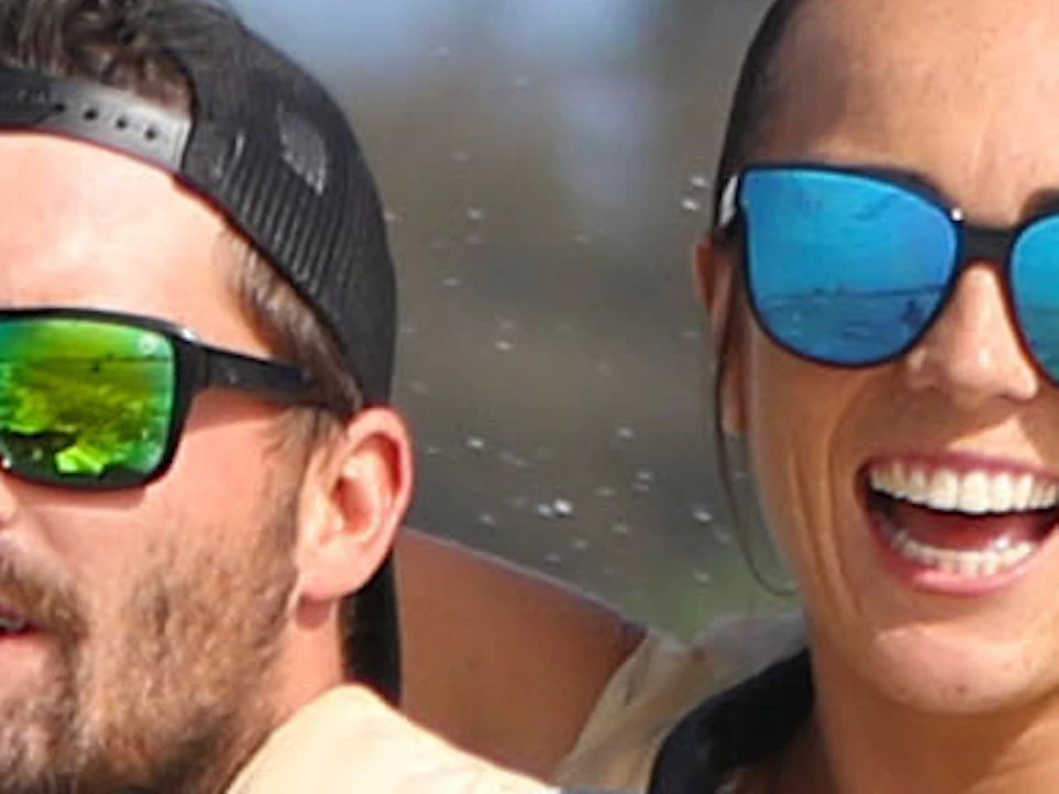 Two people in life vests riding a jet ski on water. The woman in back has arms raised. A boat and trees are in the background.
