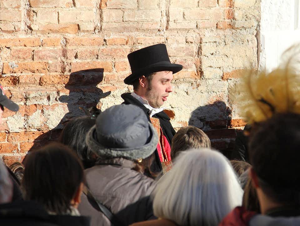 太陽に照らされたレンガの壁の前で、シルクハットをかぶった男性が集まった群衆に話しかけている。