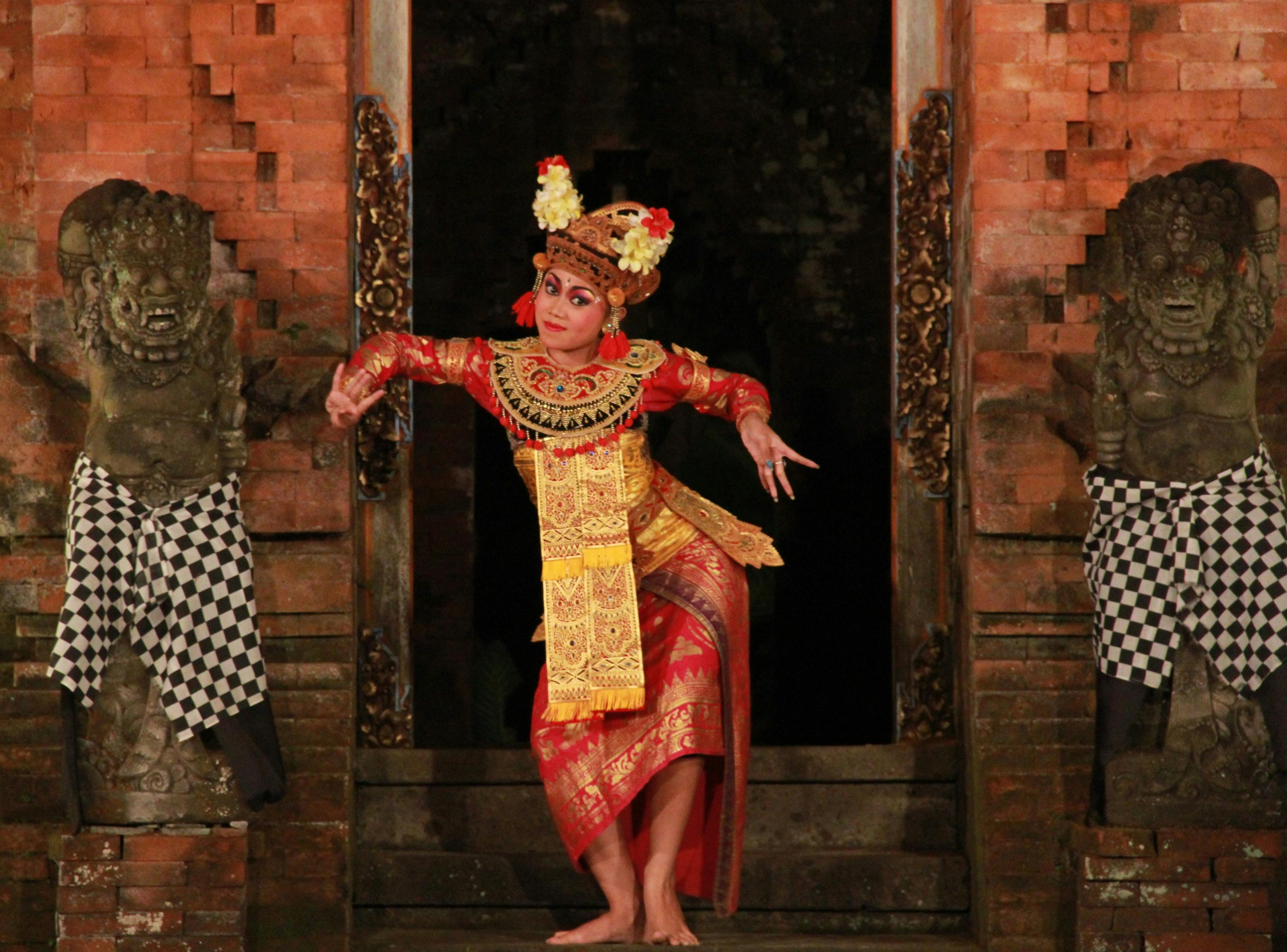 A performer in traditional Balinese attire dancing in front of an ornate temple entrance, with detailed carvings and a stone statue.