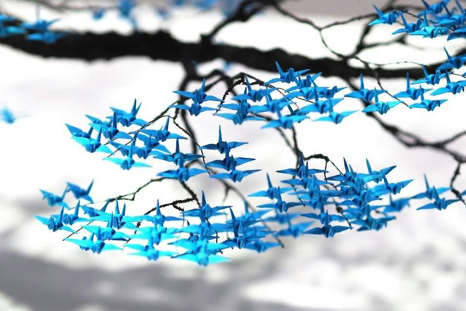 Branches adorned with numerous small blue origami cranes against a soft, blurred background.
