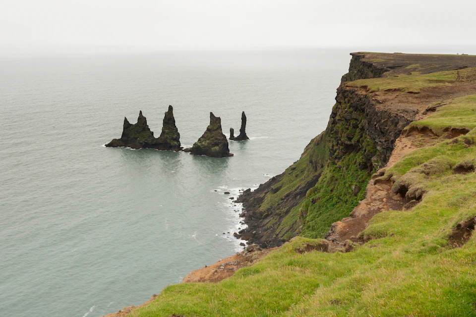 Cliffs overlooking the ocean with three jagged rock formations emerging from the water under a cloudy sky.