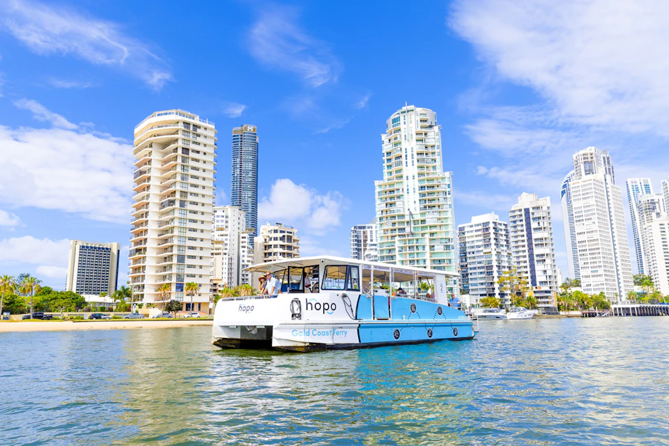 A ferry labeled 'Hopo Gold Coast Ferry' on the water with tall buildings in the background under a sunny blue sky.