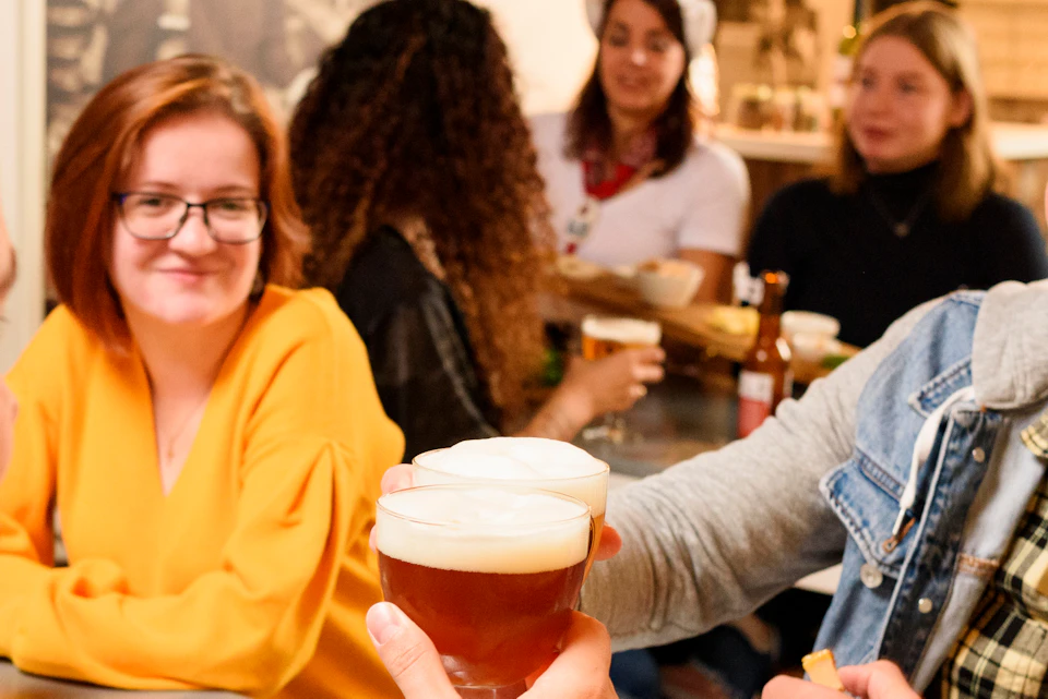 People in a bar toasting with beer; various small dishes on the table. Smiling patrons in the background.