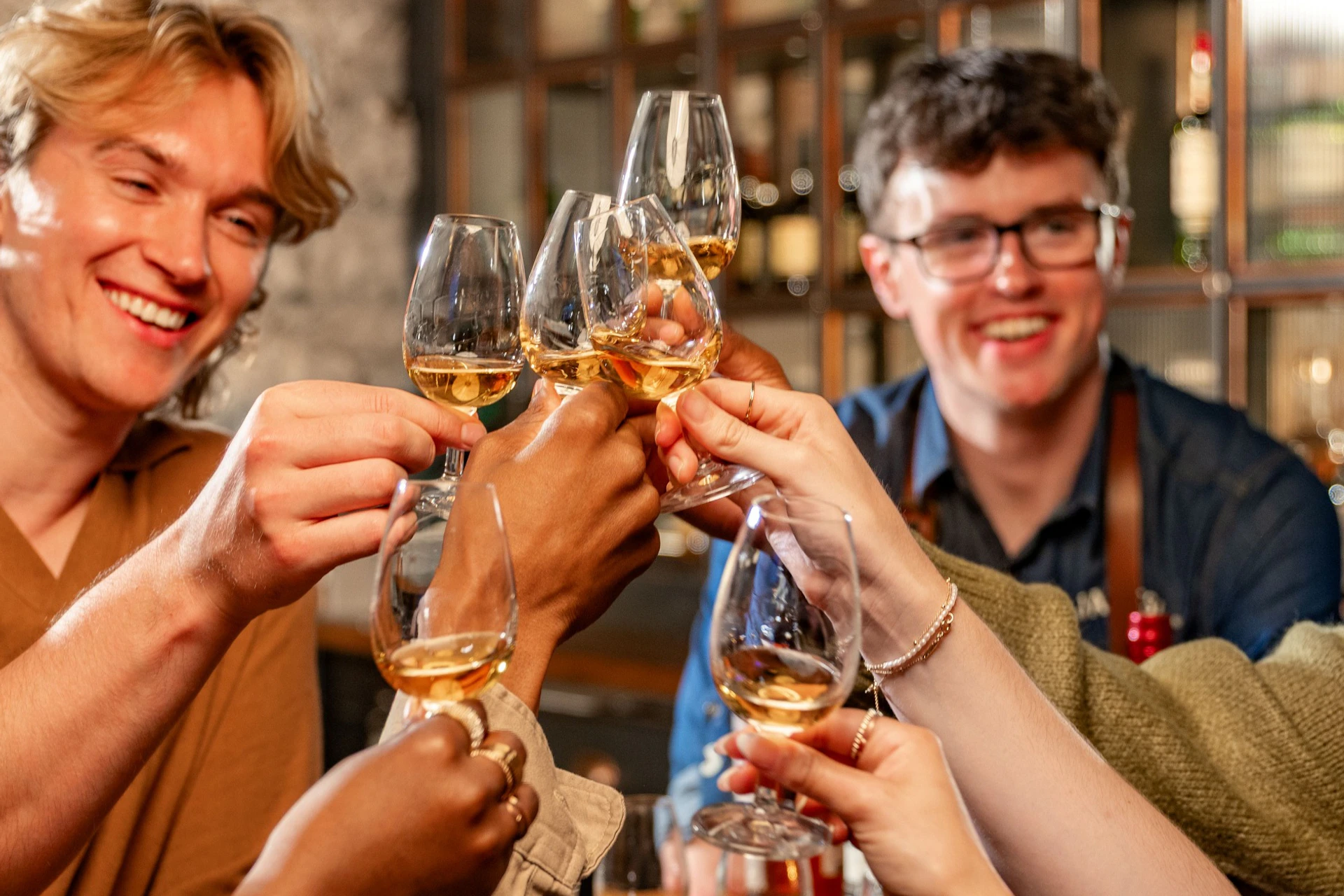 A group of friends clinking glasses at a bar, smiling and celebrating, with shelves of bottles in the background.