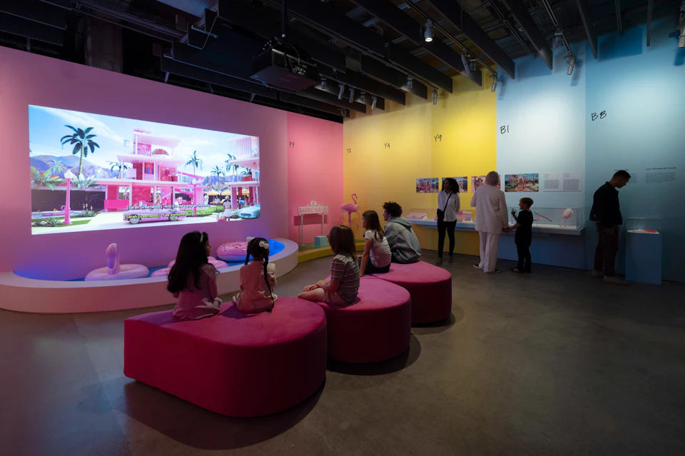 A museum exhibit with pink seating, children observing a colorful display, and adults standing by a wall exhibit.