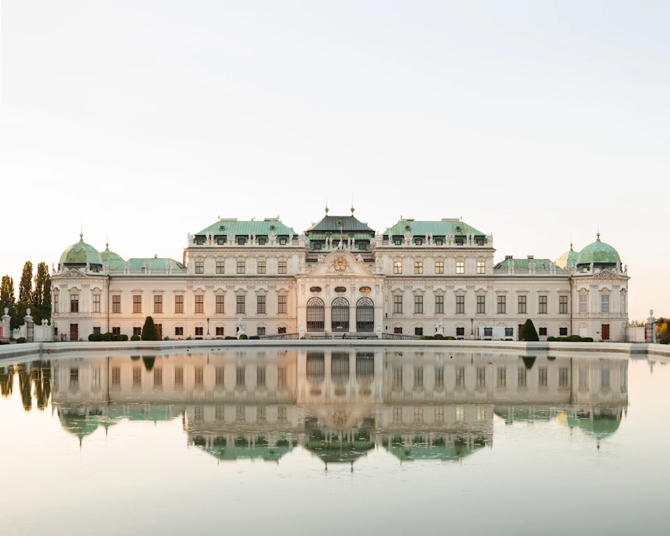 Large, ornate building with green-topped towers reflected in a calm pond, set against a clear sky.