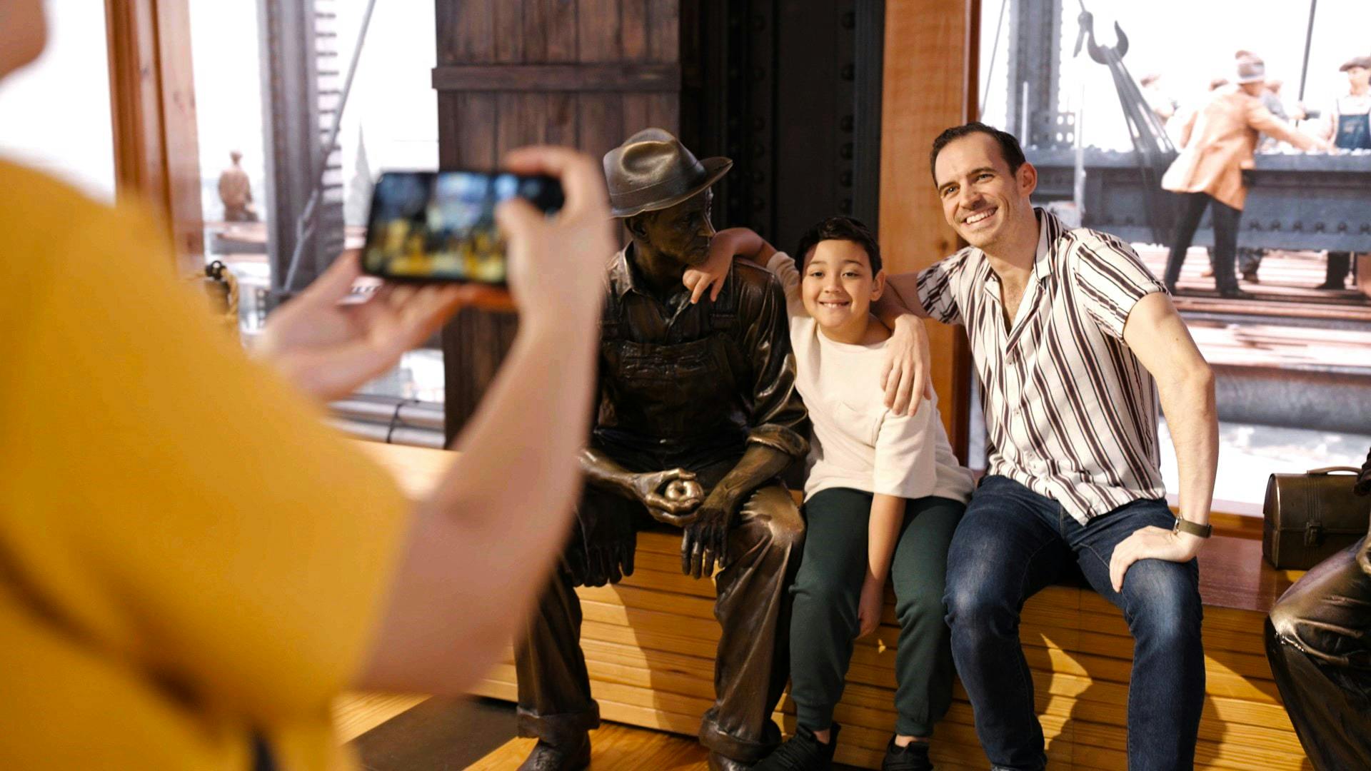 Two people posing with a seated bronze statue in a museum setting. A background features a historical photo of workers.
