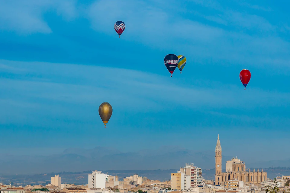 Multiple hot air balloons float in a bright blue sky above a cityscape with a prominent church tower.