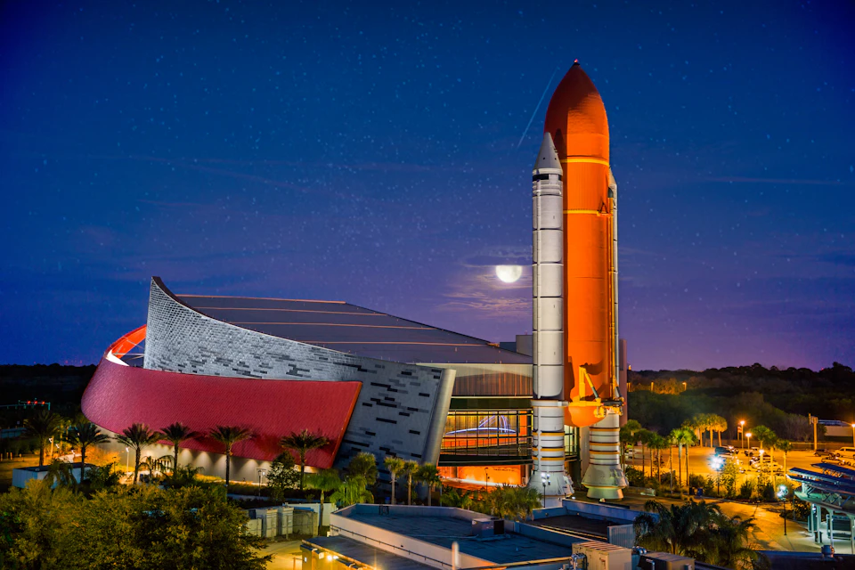 A space shuttle replica stands beside a modern building under a night sky with a visible moon, surrounded by palm trees.
