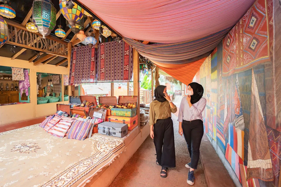 Two women wearing headscarves walk through a colorful market with fabric and lantern displays on both sides.