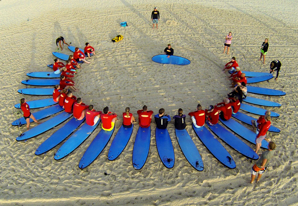 A group of people in red shirts sit on blue surfboards arranged in a circle on a sandy beach, with instructors in the center.