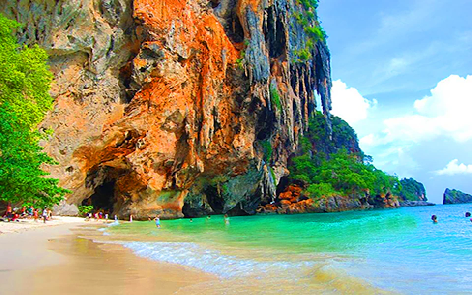 Turquoise water and sandy shore beside towering, colorful cliffs with greenery. People are swimming near the cave entrance.