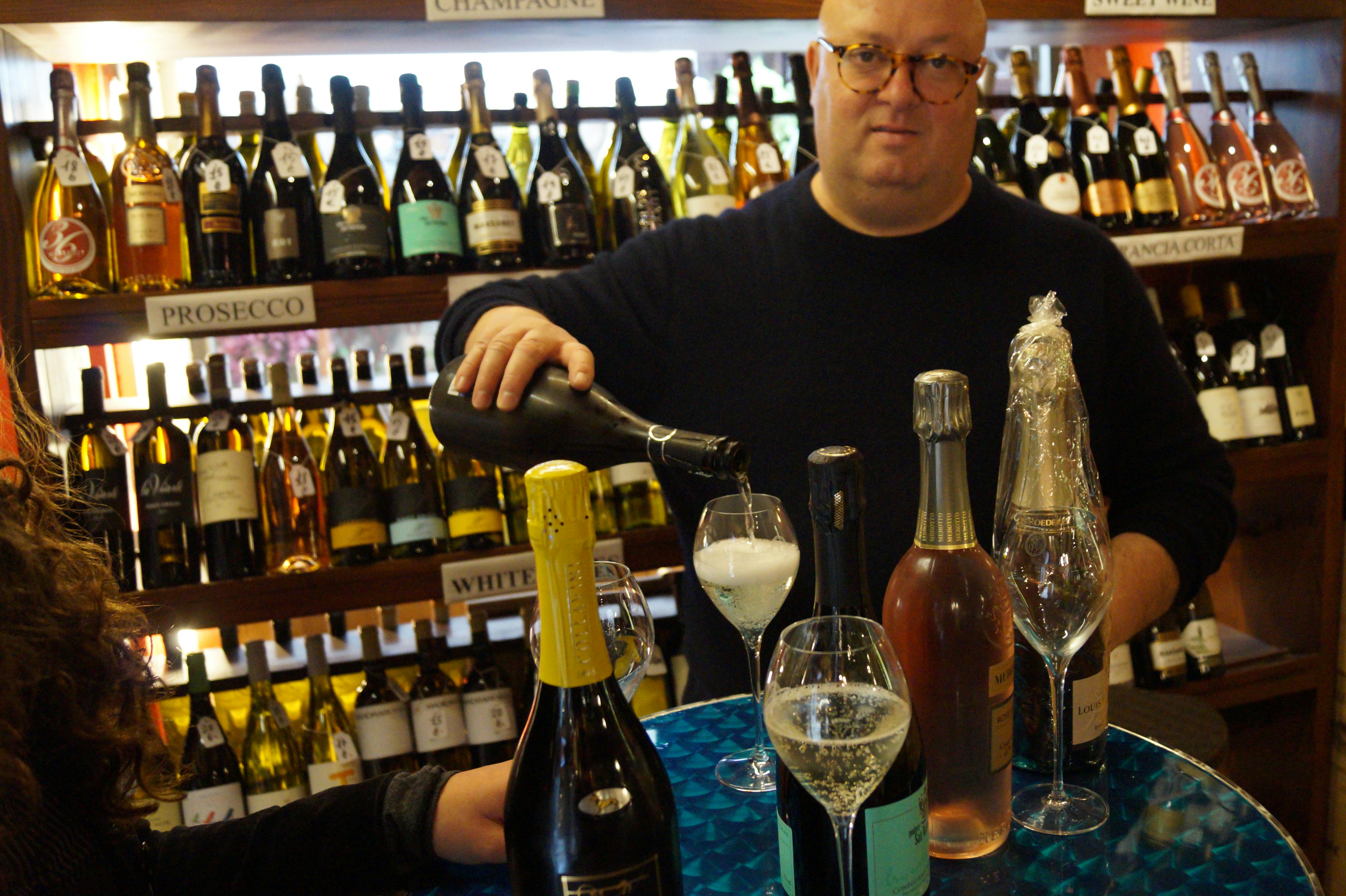 一名男子将香槟倒入桌子上的玻璃杯中，周围是商店里的各种葡萄酒瓶。
