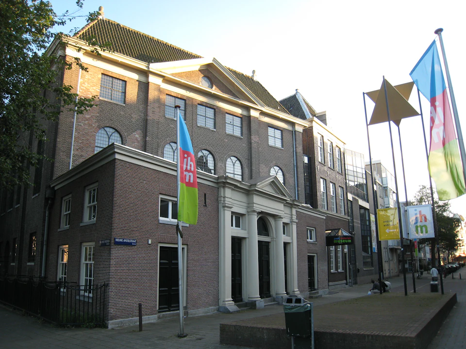Historic brick building with arched windows, a flagpole with a blue and green flag, and a street sign reading "Mari Andriessen."