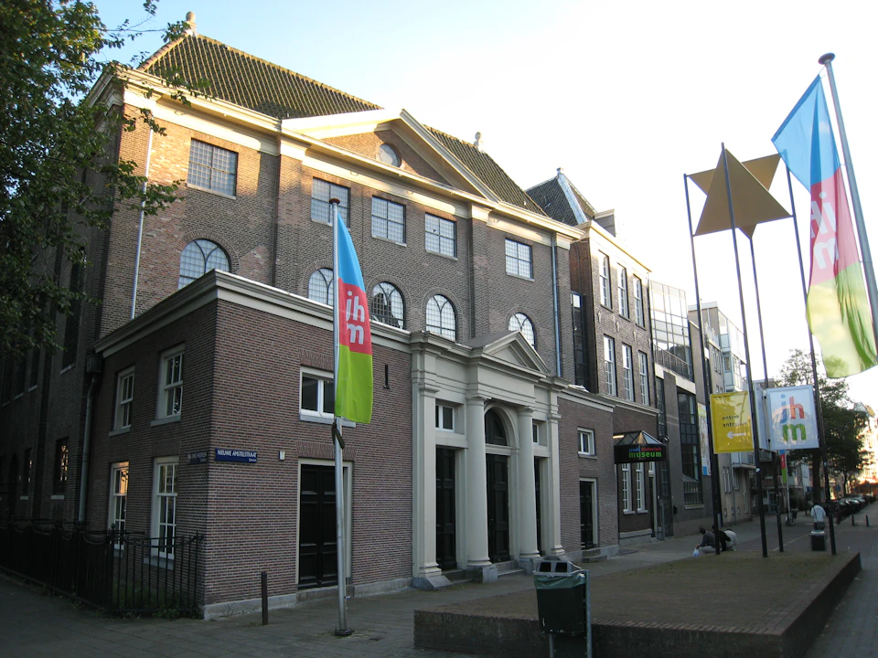Historic brick building with arched windows, a flagpole with a blue and green flag, and a street sign reading "Mari Andriessen."