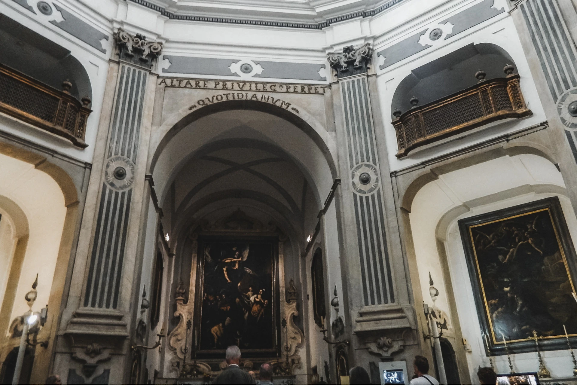 People observing artwork inside an ornate chapel with high arches, columns, and Latin inscriptions on the walls.