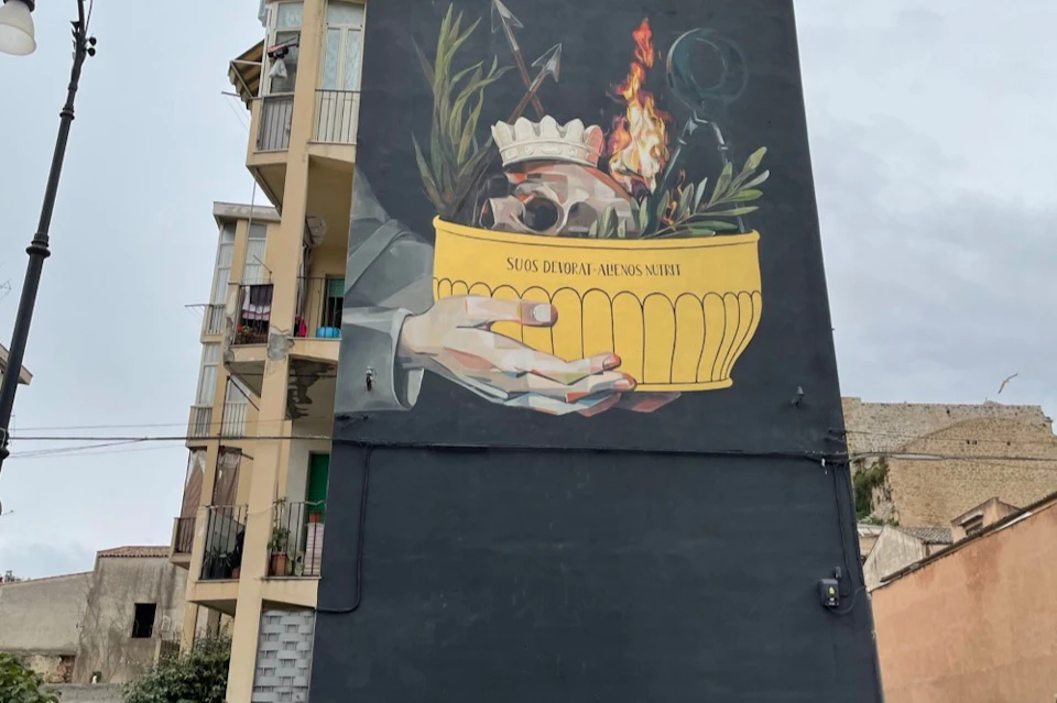 A large mural on a building wall depicts a hand holding a yellow bowl with a skull, fire, arrows, and plants.