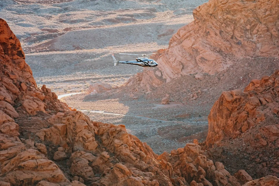 A helicopter flying over a rocky desert landscape with rugged orange cliffs and dry terrain under a sunny sky.