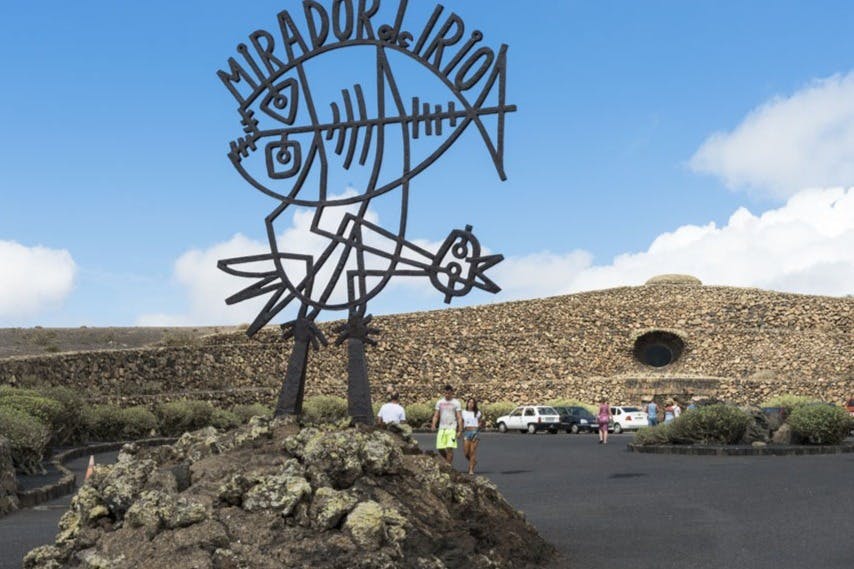 一座带有圆形窗户的石头建筑前面矗立着一座金属雕塑，上面刻着“MIRADOR DEL RIO”。 附近有人和停车。