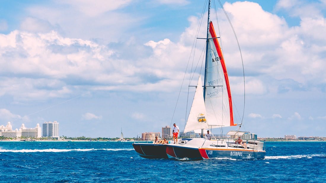 A catamaran with people aboard sailing on blue water, with a distant shoreline and buildings under a partly cloudy sky.