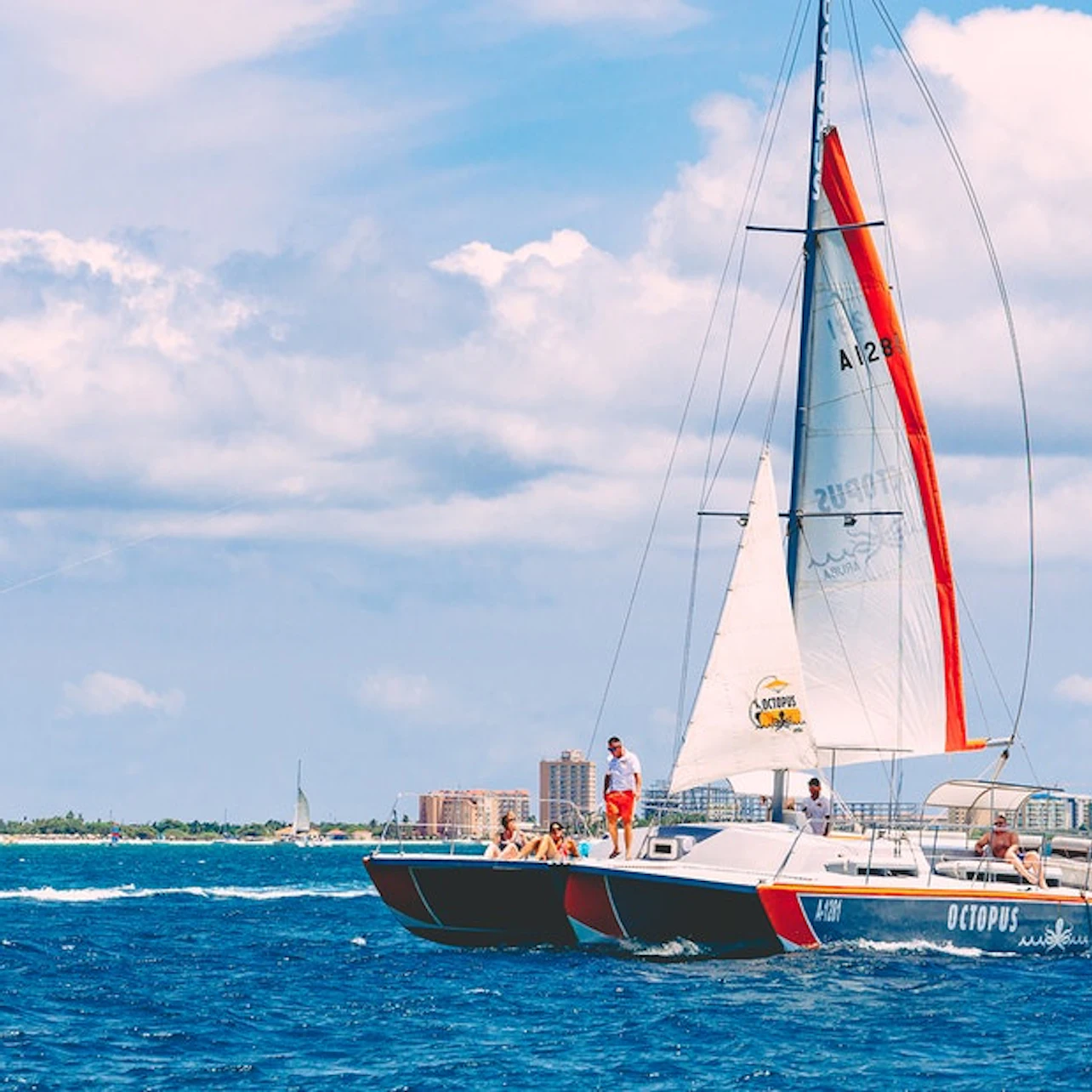 A catamaran with people aboard sailing on blue water, with a distant shoreline and buildings under a partly cloudy sky.