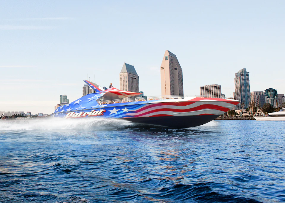A speedboat with a red, white, and blue flag design labeled "Patriot" speeds through water with a city skyline in the background.