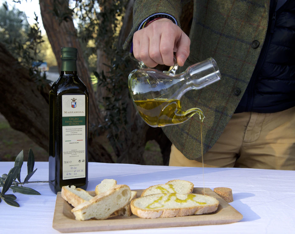 A person pouring olive oil from a glass jug onto sliced bread on a wooden board, with a bottle of olive oil nearby.