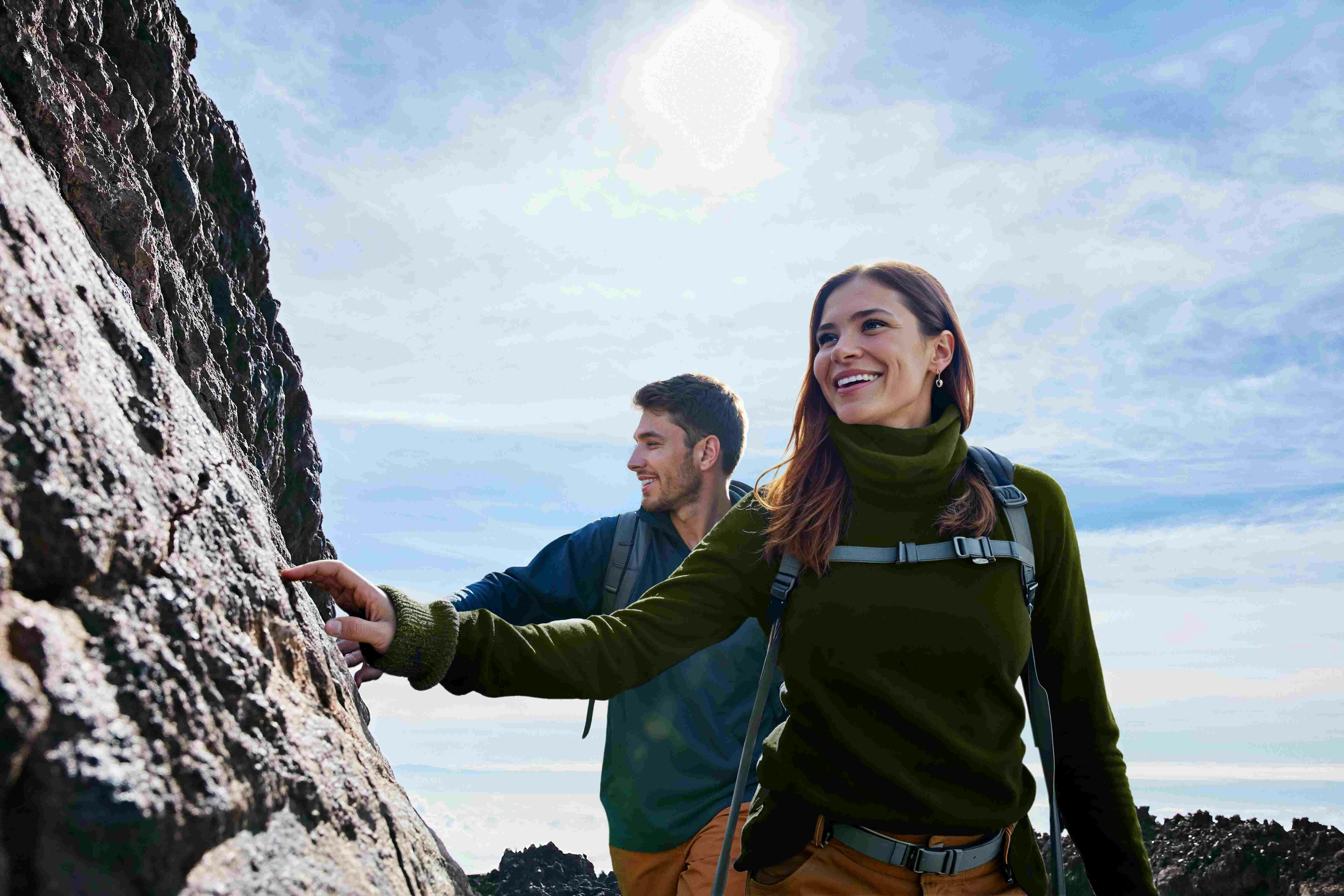 Two hikers with backpacks smiling and climbing a rocky terrain under a clear sky.