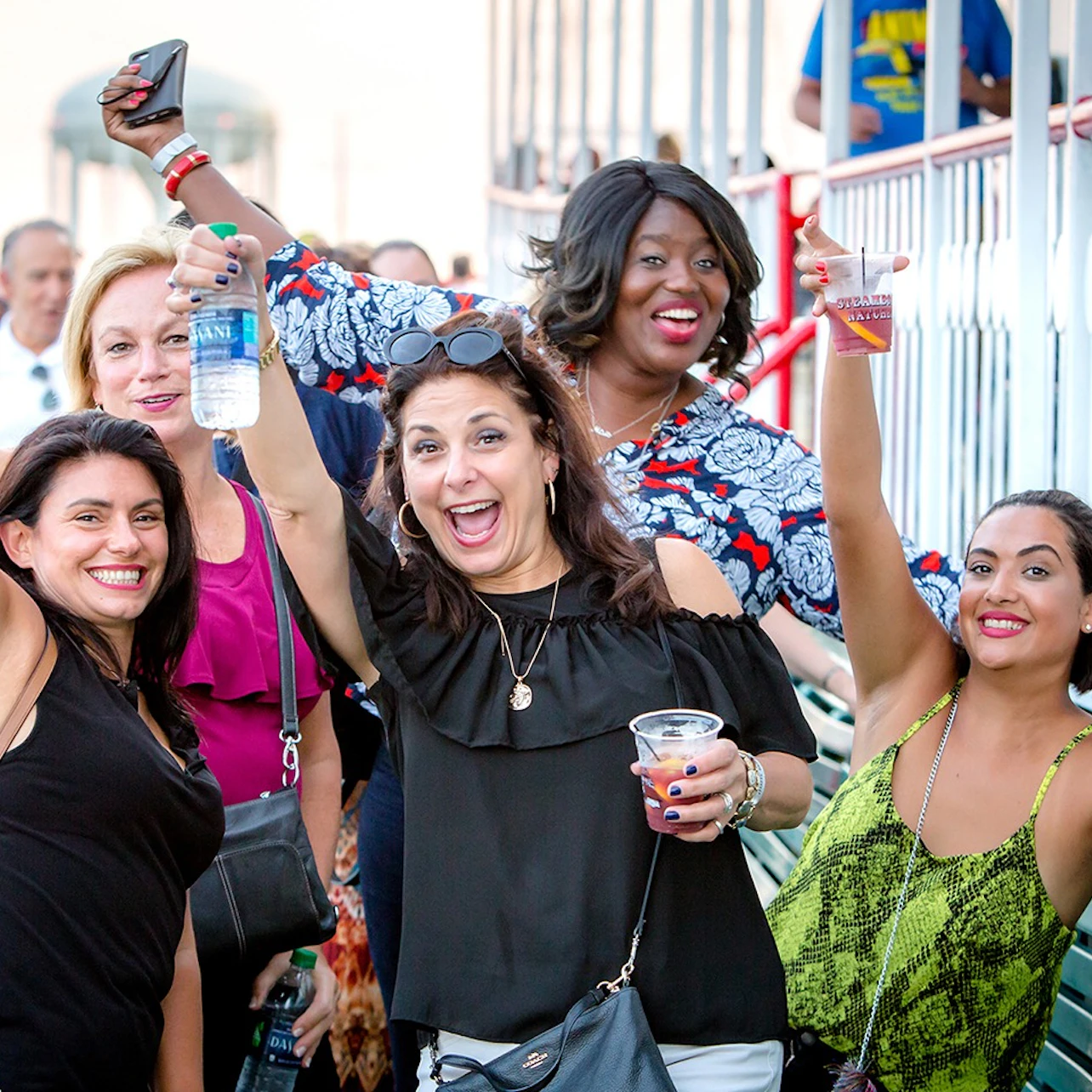 A group of people smiling and raising drinks in a celebratory manner in an outdoor setting with railings in the background.