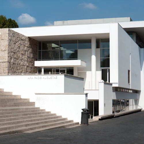Modern vit byggnad med stora glasfönster och stenaccenter, märkt "ARA PACIS AUGUSTAE." Trappsteg och träd synliga.