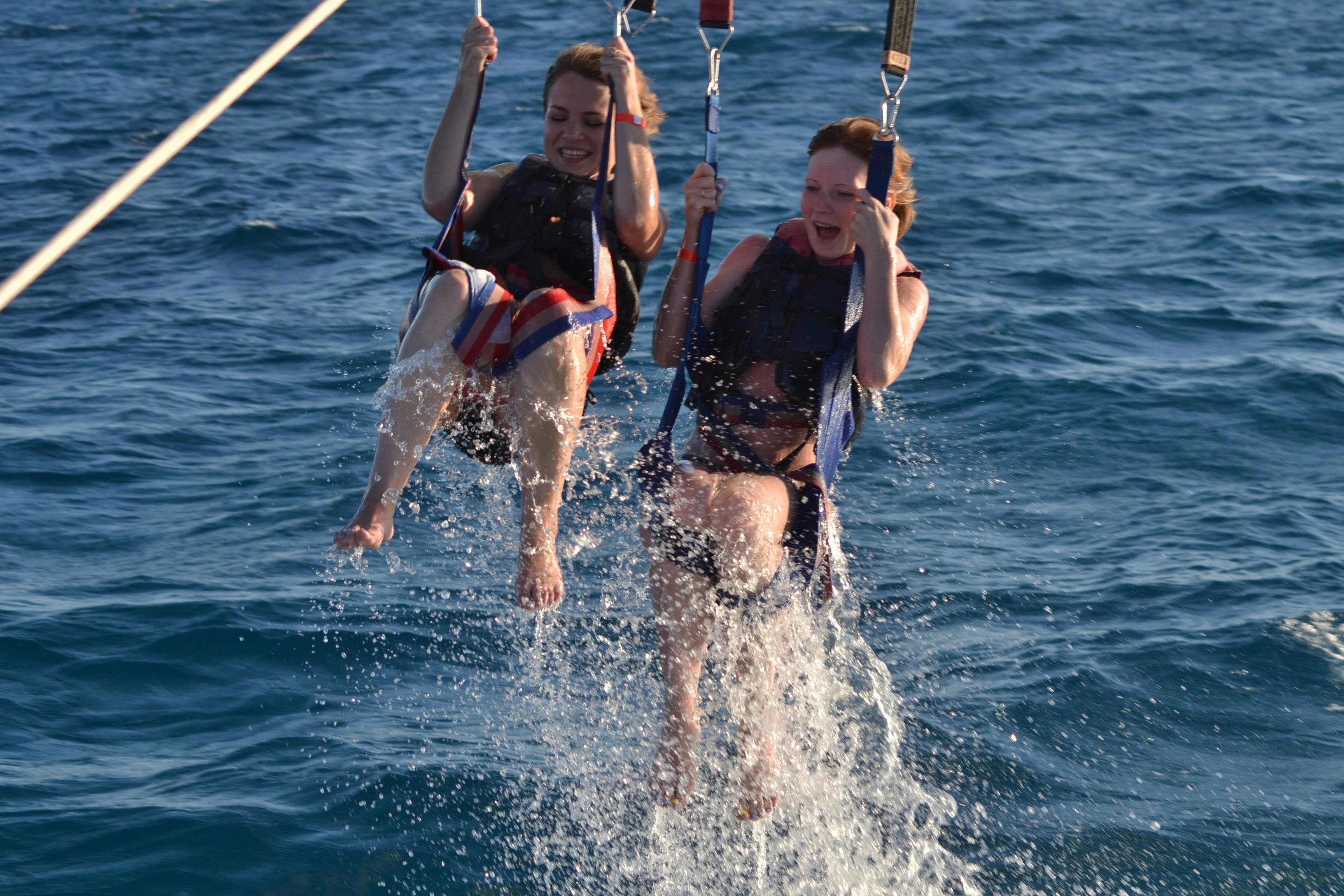 Two smiling people in life vests are parasailing, with their feet touching the water, creating splashes.