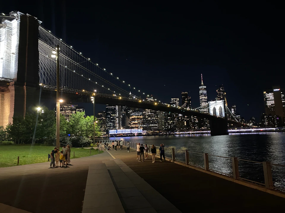 Night view of a lit-up bridge over a river with a city skyline in the background and people walking along the promenade.