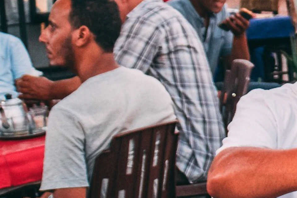 Three men sit at an outdoor table, toasting with small glasses of tea in a busy market area with vendors and stalls.